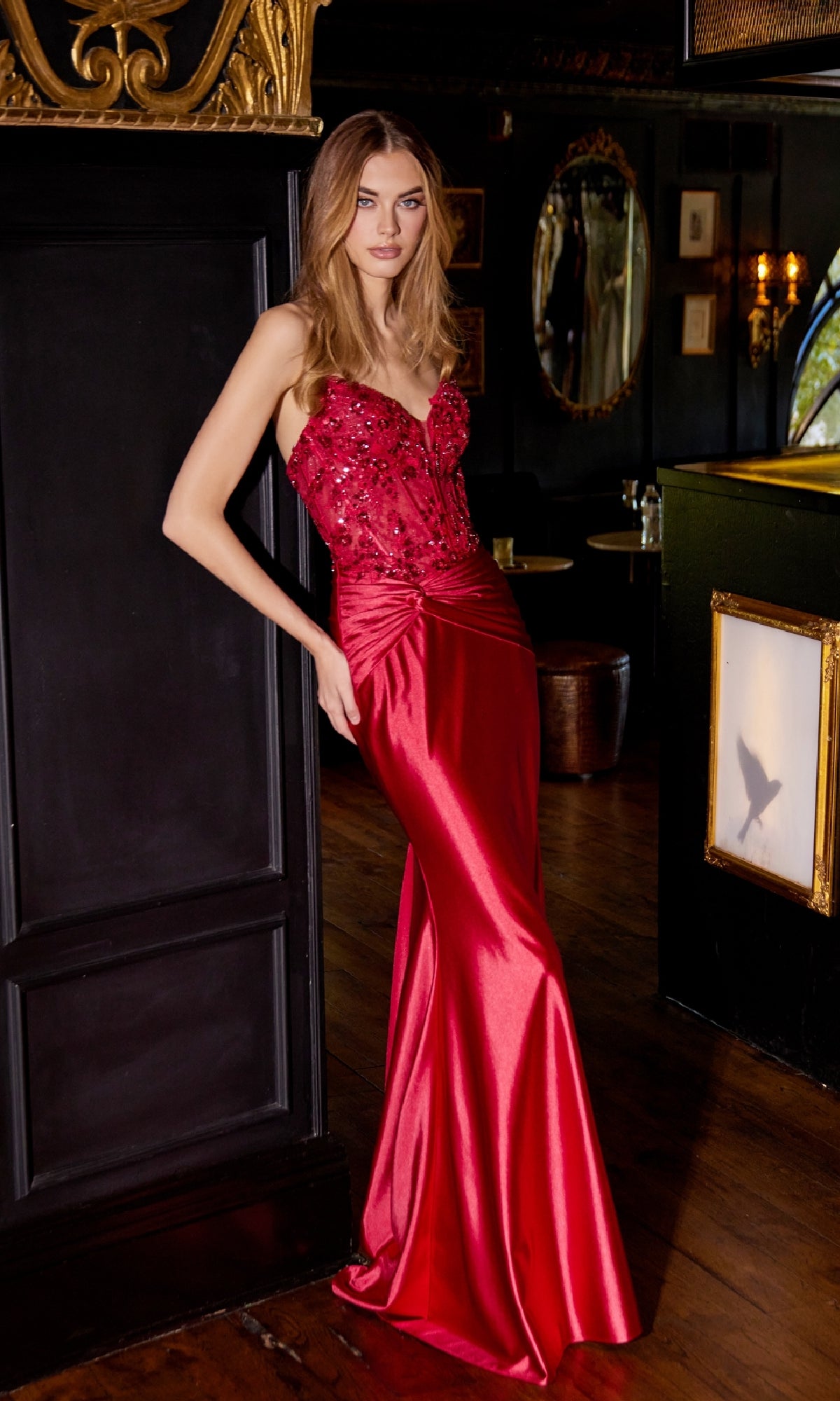 A woman stands indoors by a dark wall with ornate gold trim, wearing the Sequin-Bodice Long Prom Dress: Juliet JT2534K, featuring a strapless design and embellished corset bodice.